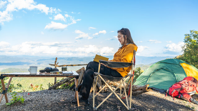 Asian woman remote working corporate business on digital tablet and smartphone while travel nature camping on mountain peak with drinking coffee. Digital nomad workation lifestyle with nature therapy