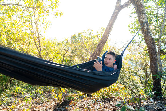 Asian man relaxing in hammock using smartphone while camping in forest enjoy outdoor active lifestyle leisure travel digital connection social media communication, nature healing on holiday vacation.