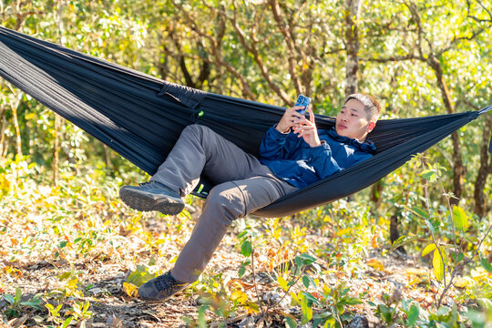 Asian man relaxing in hammock using smartphone while camping in forest enjoy outdoor active lifestyle leisure travel digital connection social media communication, nature healing on holiday vacation.