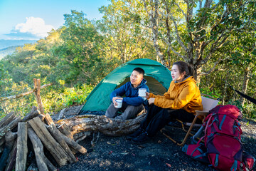 Fototapeta premium Happy Asian couple drinking hot coffee while travel nature and camping at forest mountain. Man and woman relaxing at campsite, enjoying morning beverage with outdoors active lifestyle nature therapy.