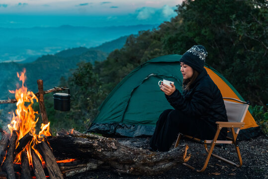 Asian woman drinking hot coffee beside campfire and tent in forest mountain campsite. People enjoy travel nature outdoor active lifestyle hiking, backpacking and wilderness recreation nature healing.