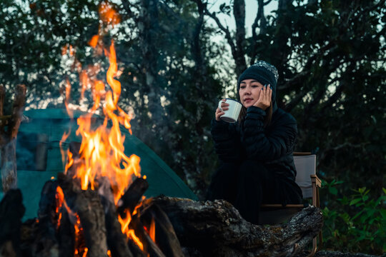 Asian woman drinking hot coffee beside campfire and tent in forest mountain campsite. People enjoy travel nature outdoor active lifestyle hiking, backpacking and wilderness recreation nature healing.