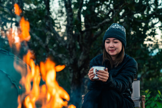 Asian woman drinking hot coffee beside campfire and tent in forest mountain campsite. People enjoy travel nature outdoor active lifestyle hiking, backpacking and wilderness recreation nature healing.