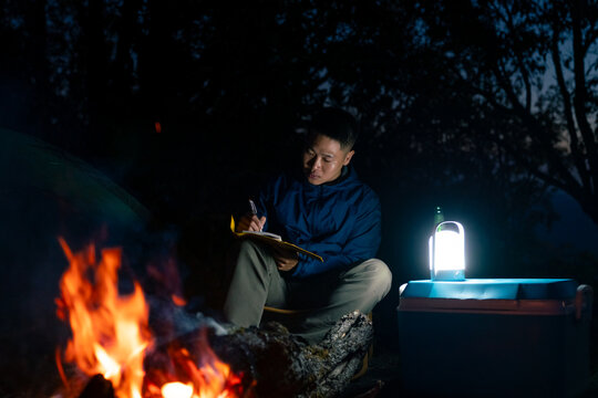 Asian man writing travel journal and drinking hot coffee beside campfire and tent in forest mountain. People relax and enjoy travel outdoor active lifestyle hiking, backpacking and nature healing.