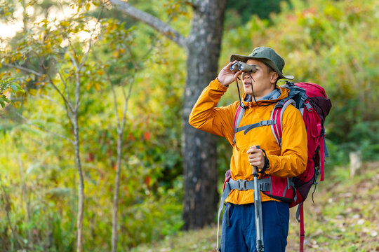 Asian man using binoculars while hiking in forest with backpack and trekking pole. Male traveler enjoy outdoor active lifestyle solo travel, adventure journey and wildlife observation nature therapy.