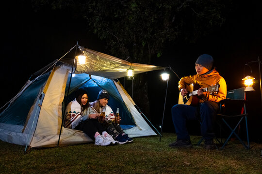 Group of Adult friends camping on mountain, drinking beer and playing guitar together by tent. Man and woman enjoy outdoor active lifestyle campsite leisure and friendship travel on holiday vacation.