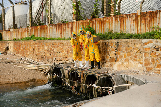 Environmental field workers in protective hazmat suits collect water samples from floodwater beside a drainage channel, conducting contamination assessment and water quality monitoring.