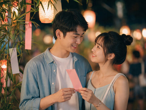 Young Asian couple sharing a pink wish card beside bamboo at a lantern festival, affectionate eye contact and soft night bokeh for Qixi romance.