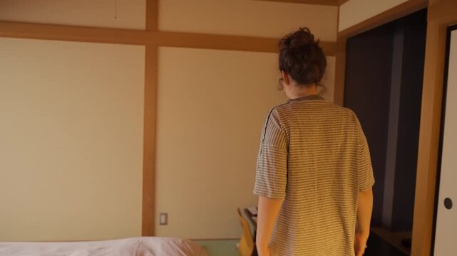 Happy woman exploring a traditional Japanese ryokan room in Japan. Walking towards the window to enjoy the morning light during her vacation. Simple room with Tatami mat and futon bed