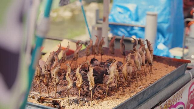 Delicious ayu fish on skewers grilling over hot charcoal at an outdoor street food stall. Traditional Japanese summer delicacy, shioyaki, being cooked at a festival market in Japan