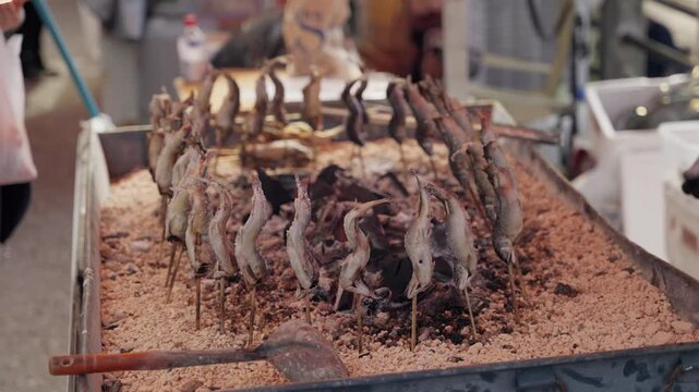 Ayu, or sweetfish, grilling on skewers over hot charcoal embers in a traditional Japanese irori style sand pit, a popular delicacy and street food at a bustling market in Takayama, Japan