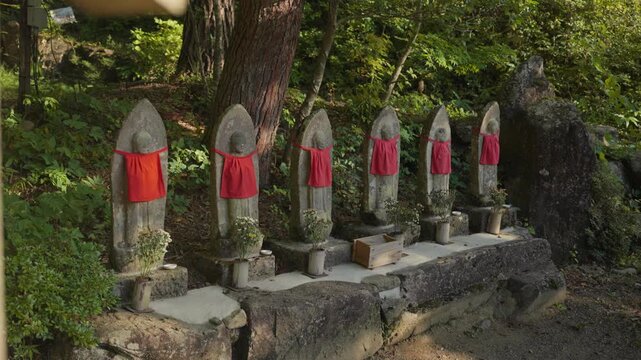 Row of ancient Jizo statues, protectors of children and travelers, wearing traditional red bibs in a tranquil forest setting at a Buddhist temple in Hokkaido, Japan, surrounded by lush green nature