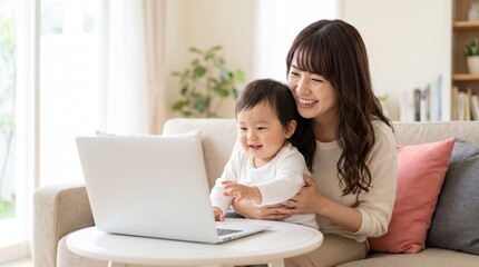 Fototapeta premium Happy Japanese mother sitting on living room sofa with her baby daughter, smiling while using a laptop computer for online communication at home