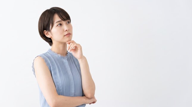 Thoughtful young Asian woman looking up with hand on chin while thinking or worrying, standing against a white studio background with copy space