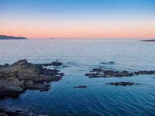 Fototapeta premium Petite crique rocheuse au crépuscule avec eau calme et transparente, paysage marin serein illuminé par des teintes roses et bleues douces