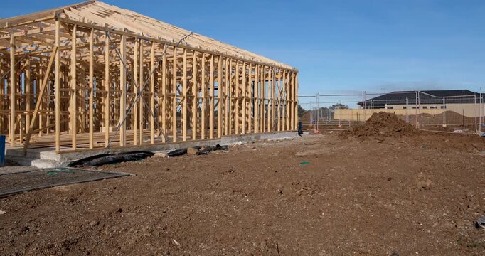 a residential house frame under construction, showing timber wall studs and roof trusses built on a concrete slab foundation across an open dirt building lot in an outer suburb of Melbourne, Australia