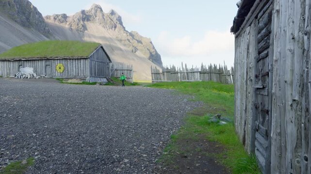 Old Viking village film set at Stokksnes, Iceland, featuring turf houses and wooden fences against the iconic Vestrahorn mountain range. A lone hiker explores this remote, historic landscape.