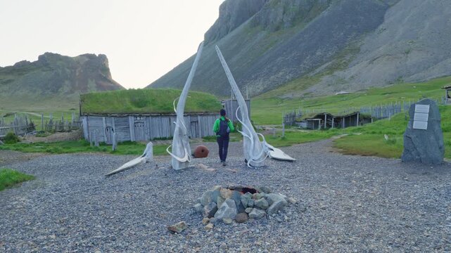 A lone hiker explores the atmospheric old Viking village film set on Iceland. Features traditional grass-roofed huts, ancient-looking whale bones, and a rocky mountain landscape under soft light.
