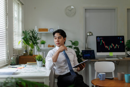 Young male investor holding a digital tablet, looking thoughtfully while planning stock market investments in a modern home office with trading charts on computer screens