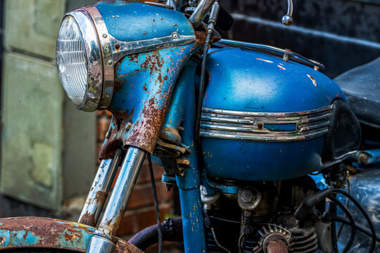 Vintage rusty blue motorcycle parked outside a garage, detail view of classic engine, rusted headlight, fuel tank, and patinated metal bodywork.
