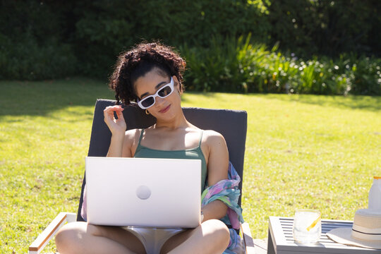 Woman sitting on cushioned lounge chair in backyard, using laptop and wearing colorful wrap