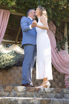 Couple standing on low stone ledge under wooden pergola, holding hands, wearing suit and lace dress