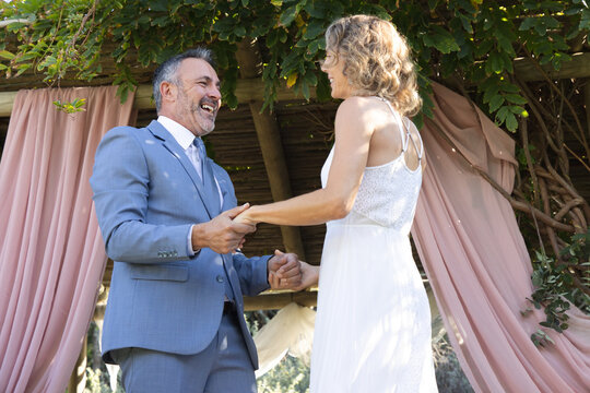 Couple standing under wood arbor pink drapes garden holding hands groom blue suit bride white dress