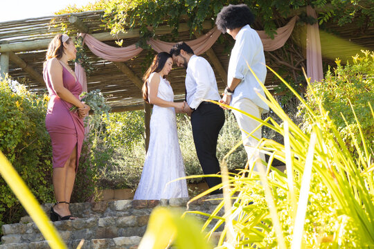 Diverse wedding party showing African American bride in gown and groom holding hands under pergola