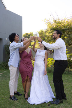 Diverse wedding party raising champagne flutes on garden lawn, bride wearing white gown