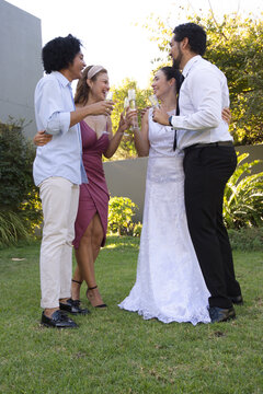 Diverse couples toasting and laughing in garden holding champagne flutes, wedding gown and tie