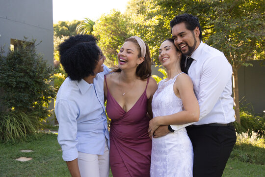 Diverse friends laughing and embracing on backyard lawn, wearing white lace dress and black necktie