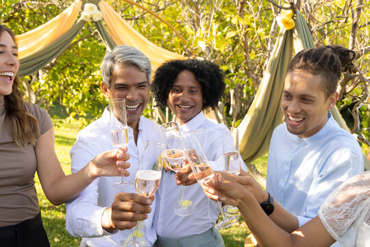 Diverse friends raising champagne flutes, laughing and toasting in garden under fabric drapery