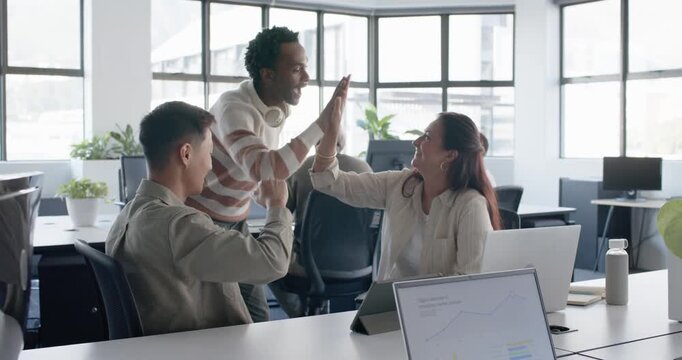 Diverse team in office high-fiving, pointing and using tablet and laptop reviewing charts