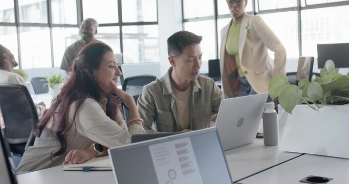 Diverse coworkers leaning over laptop showing charts, reviewing at open-plan desk and taking notes