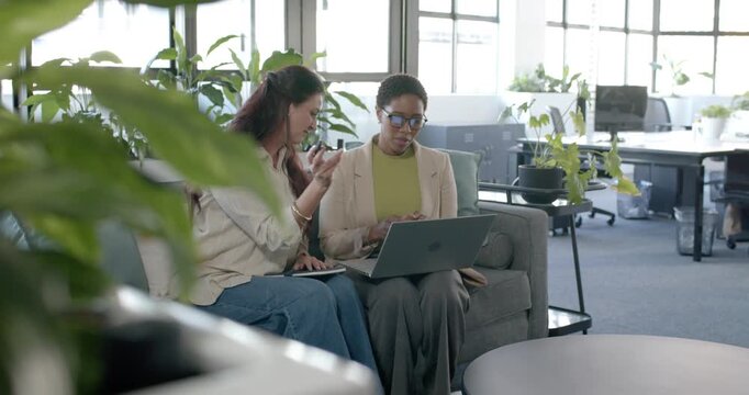 Female coworkers joining, showing phone and passing pen while comparing laptop at office for work