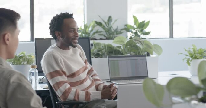 Diverse team watching coworker laugh as woman walking up to desk leaning reviewing laptop