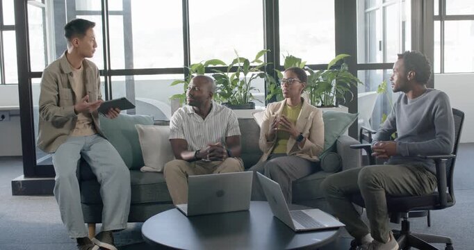 Diverse coworkers hearing Asian man tapping tablet, talking in lounge with laptops, seeking input