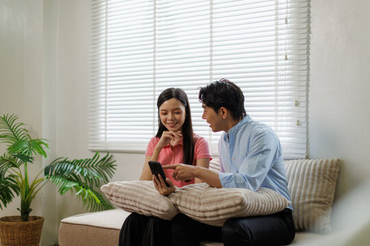 Young asian couple relaxing on a living room sofa, happily using a smartphone together, pointing at the screen and sharing content, embodying connection and modern lifestyle