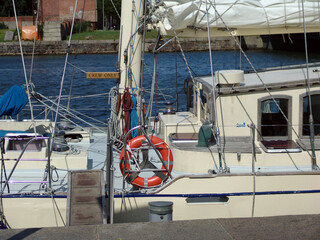 Close‑up of a classic sailing yacht moored in a marina, featuring rigging, life ring, deck hardware, cabin windows, and nautical equipment along a waterfront harbor. © Martins
