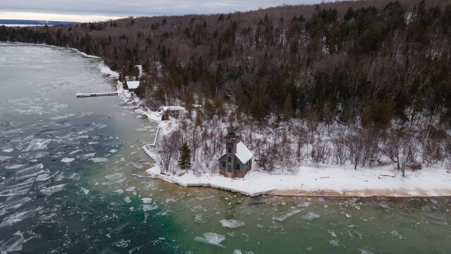 Aerial view of Grand Island Lighthouse situated on a snow-covered shoreline with icy waters and dense forest in Munising, Michigan, United States.