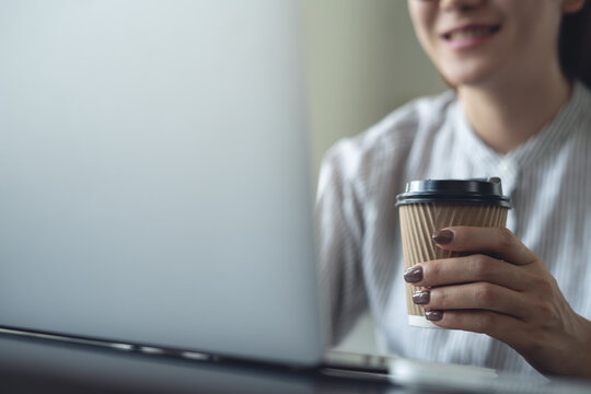 Asian woman working remotely using laptop, hand holding a coffee cup, work at home, business casual, freelance lifestyle concept