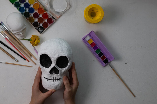 Unpainted paper-mache Day of the Dead skull held over a white craft desk, pencil outlines visible, showcasing handmade, DIY skull decoration materials and a soft daylight studio setup.