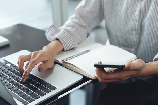 Business woman using smartphone while working on laptop, searching online content, working remotely at modern workspace