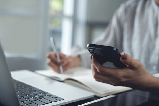 Close up of business woman using smartphone and writing on notebook, planning new business project during working on laptop at home office