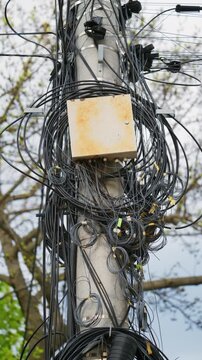 Tangled mess of wires and cables on an electricity pole, with a junction box, against a backdrop of trees.