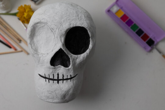 Unfinished handmade Day of the Dead skull sculpture on a craft table; pencil outlines reveal floral patterns as creators prepare decoration with colors and tools in a warm studio.