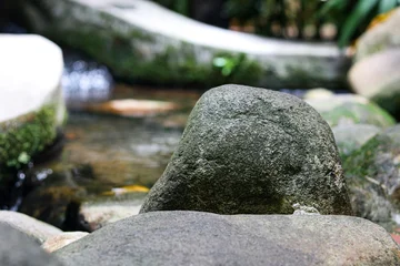 Fotobehang Rustgevend A smooth, moss-covered stone sits nestled among larger rocks along the edge of a shallow, peaceful stream.  © Ink N Propeller
