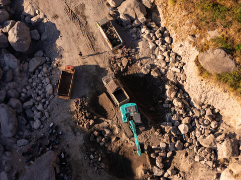 Aerial view of an active stone quarry with an excavator loading rocks into a dump truck as workers stand nearby in Klaten, Central Java, Indonesia.