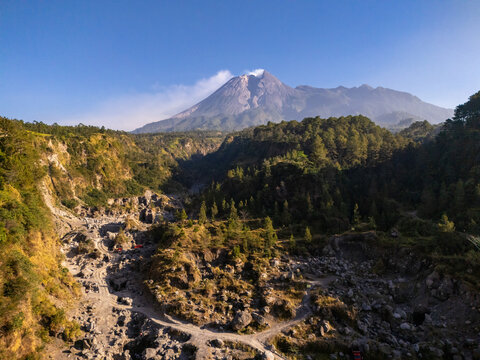 Aerial view of Mount Merapi with its volcanic plume rising above a rocky riverbed and lush green forested slopes under a clear blue sky in Klaten, Central Java, Indonesia.