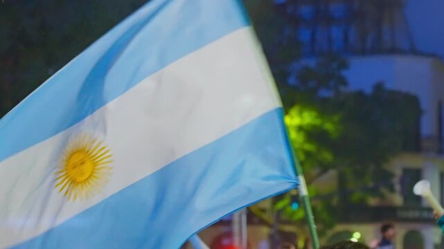 An Argentina flag waves close up in front of a tree lined Buenos Aires plaza at night, someone climbing a lamppost in the background, on July 19, 2026.